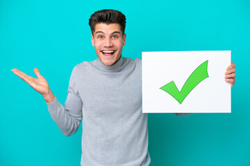 Young handsome caucasian man isolated on blue bakcground holding a placard with text Green check mark icon with surprised expression