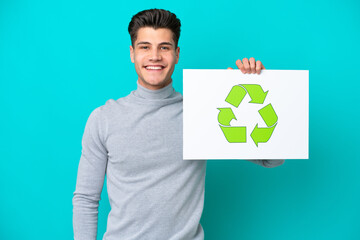 Young handsome caucasian man isolated on blue bakcground holding a placard with recycle icon with happy expression