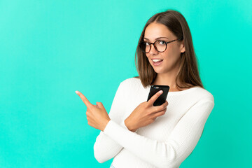 Young woman over isolated blue background using mobile phone and pointing back