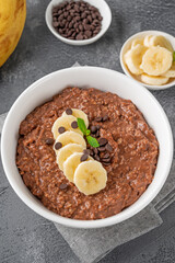 Chocolate oatmeal porridge with banana and chocolate chips on top in a white bowl. Healthy breakfast. Copy space.