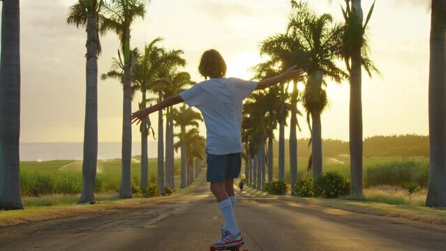Happy care free boy skateboarding down street at sunset with hands up in air. Palm trees by a blue sky. Driving through the sunny Beverly Hills. Los Angeles, California.
