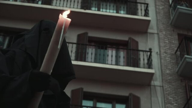 Close-up shot of the melting candle and in the background the covered face of a worshipper in a black tunic. Holy Week in Spain.