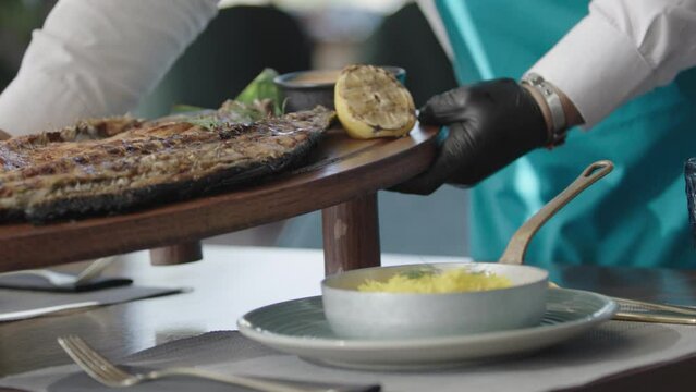 Waiter serving a grilled fish with lemon and sauce  on the table. 2K MXF.