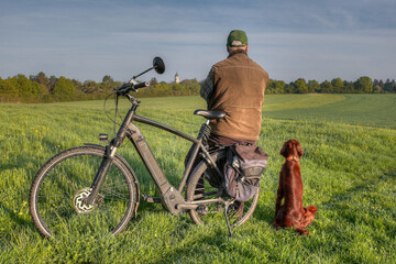 A man sits on the luggage rack of his bicycle in the sun on a green meadow. His Irish Setter dog is...