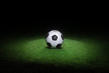 Black and white leather football ball laying on fresh green grass highlighted by spotlight.