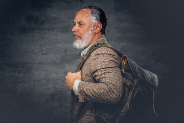 Portrait of gray haired old man dressed in beige suit carrying backpack against dark background.