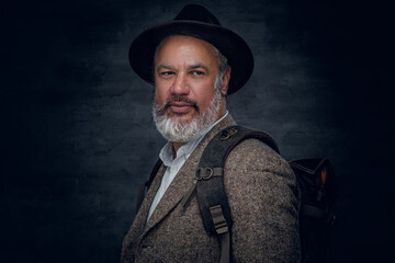 Studio shot of stylish grandfather dressed in beige suit and hat carrying backpack.