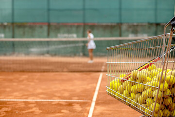 Lots of tennis balls in a metal basket on tennis court on background of tennis player watering the court