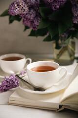 A cup of green tea against the background of a spring bouquet of lilacs on a textured gray background.Romantic composition with books and candles. Spring tea drink. Side view. Place to copy.