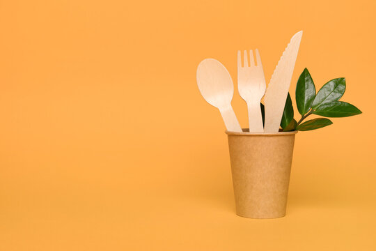 Natural wooden bamboo cutlery set in paper cup - biodegradable and compostable cutlery and tableware concept. Bamboo cutlery on orange background with copy space. Environmental conservation concept