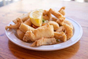 Fried cuttlefish strips, called Chocos Fritos in Spanish. It is served on a white plate on a wooden table. Spanish food concept.