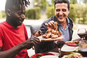 Happi multi-ethnic friends sitting at farmhouse table - an African guy hands the grilled chicken wings tray to a mid aged man - conviviality and friendship lifestyle concept - Powered by Adobe