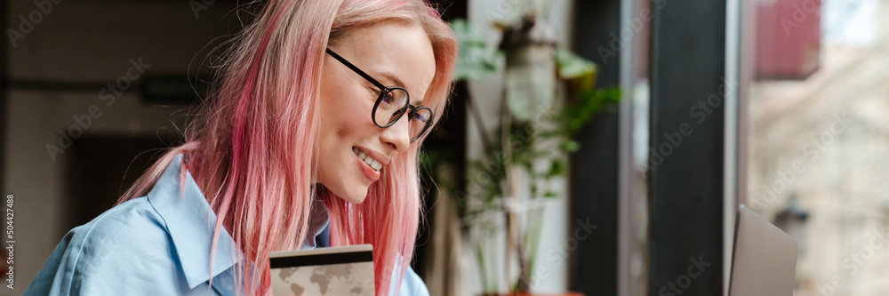 Wall mural Young woman with pink hair using laptop and credit card in cafe