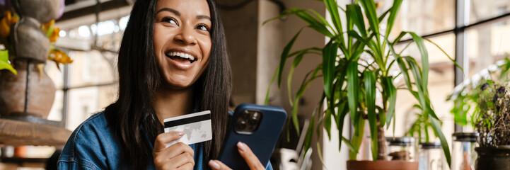 Black smiling woman using cellphone and credit card in cafe