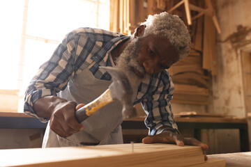 African american mature carpenter using hammer on nail in plank at workshop