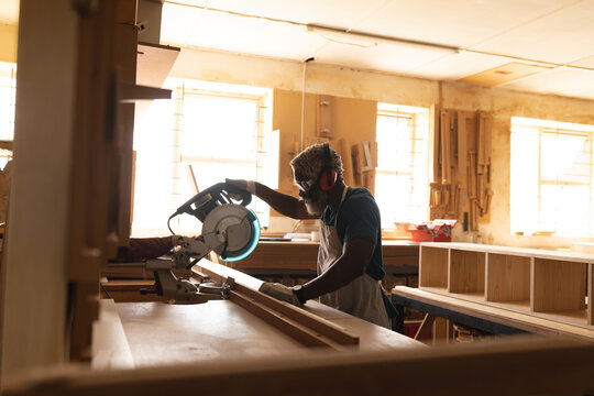 African american mature carpenter in protective eyewear and earmuffs using circular saw at workshop