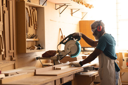 African american mature carpenter in protective eyewear and earmuffs using circular saw in workshop