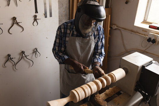 African american mature carpenter in safety face shield shaping and carving wood at workshop