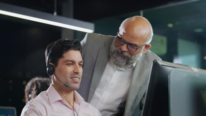 A modern Indian Asian attractive smiling customer support care male or man employee wearing headset mic working on a computer while interacting or talking to senior boss. Business, Finance concept