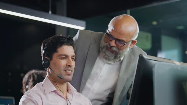A modern Indian Asian attractive smiling customer support care male or man employee wearing headset mic working on a computer while interacting or talking to senior boss. Business, Finance concept