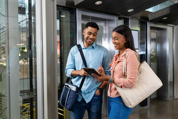 Smiling biracial businesswoman sharing tablet pc with african american businessman against elevator