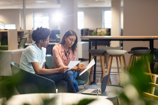 Confident female and male biracial colleagues discussing over document at modern workplace
