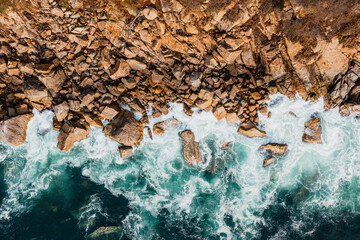 overhead shot of ocean waves crashing against the rocky shoreline in shades of blue-green and...