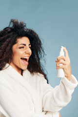 Enjoyed happy curly Latin lady in bathrobe with toner bottle in hand spraying on face posing...