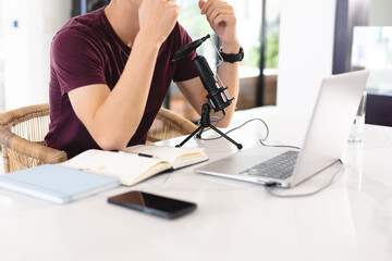 Midsection of asian teenage boy with microphone, laptop and books on table podcasting at home