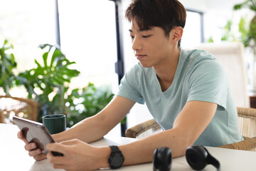 Asian teenage boy wearing blue t-shirt using digital tablet on table while sitting at home