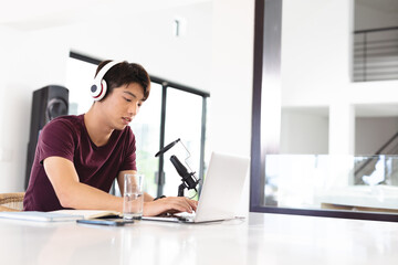 Asian teenage boy wearing headphones with microphone on table using laptop while podcasting at home