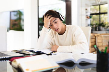 Stressed asian teenage boy wearing headphones with head in hand studying on table at home