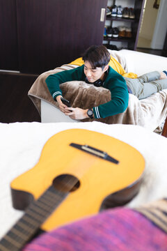 High angle view of smiling asian teenage boy using smart phone while lying over sofa in living room