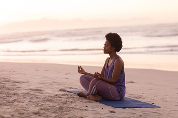 African american mature woman meditating while sitting on mat against sea and clear sky at sunset - obrazy, fototapety, plakaty