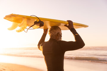 African american senior man carrying surfboard on head while standing at beach against clear sky