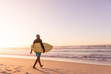 African american senior man with surfboard walking at beach against clear sky at sunset, copy space