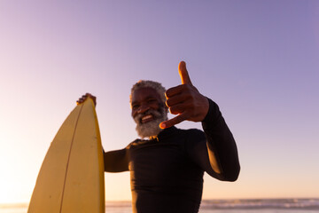 Portrait of bearded african american senior man with surfboard gesturing at beach against clear sky