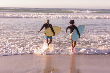 Rear view of african american couple with surfboards running amidst waves in sea at sunset
