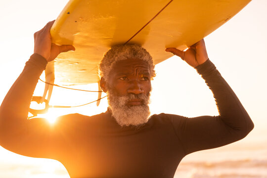 Bearded african american senior man carrying surfboard on head at beach against clear sky at sunset