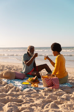 African american couple having juice while sitting with food on blanket at beach against clear sky