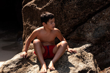 caucasian boy in red swimming trunks sit resting in a rock
