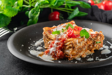 A portion of meat lasagna with bolognese sauce and mozzarella cheese on a black plate, close up