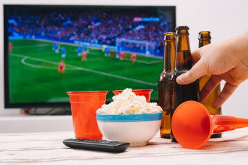 A beer with popcorn and trumpets to cheer on his football team, in front of the TV screen showing a...