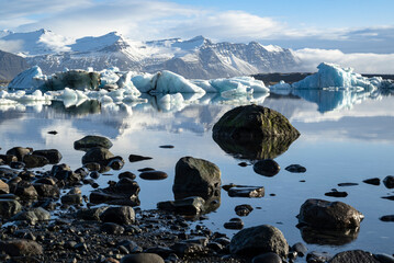 Shore of Jökulsárlón glacier lagoon with black rocks and boulders reflected in the water and floating icebergs in the distance, Iceland, Vatnajökull National Park