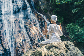 A young woman in white clothes and a turban sits on a rock near a waterfall, practices yoga and meditates