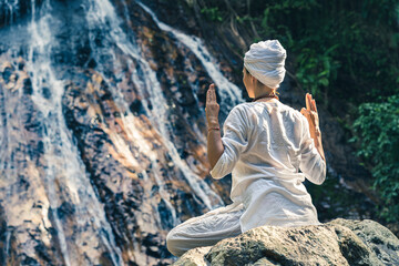 A young woman in white clothes and a turban sits on a rock near a waterfall, practices yoga and...