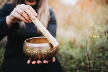 Black dressed woman outside holding and playing a tibetan singing bowl with a wooden stock. Selective focus