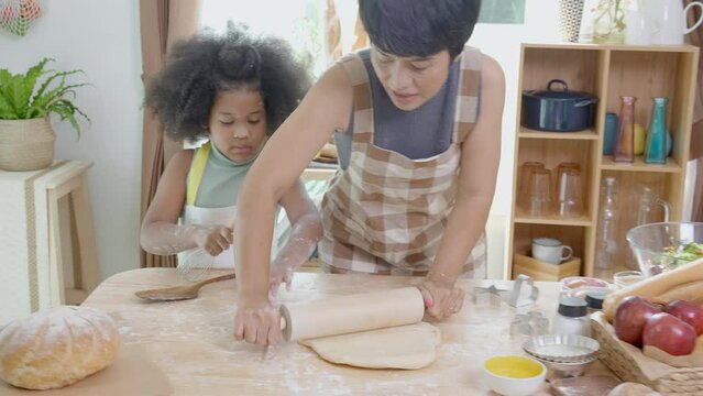 African America family with mother wearing apron rolling thresh flour for cooking with daughter together in the kitchen at home, parent and little child preparing food with fun and playful.