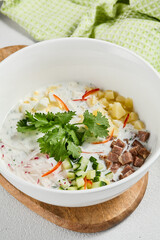 Traditional russian soups - okroshka with fresh vegetables, meat and kefir. Cold summer soup on white table. Aesthetic composition with traditional russian food and spices, textile