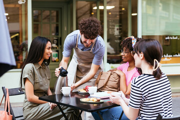 Waiter serving coffee for diverse women in summer cafe